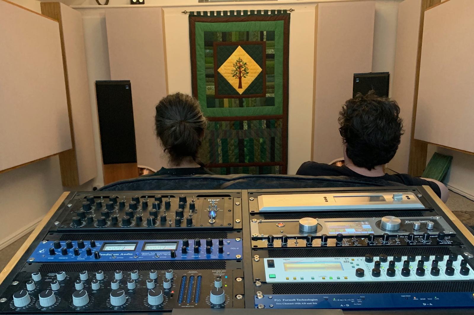 Two people seen from behind at the mastering desk, facing the monitors during a session